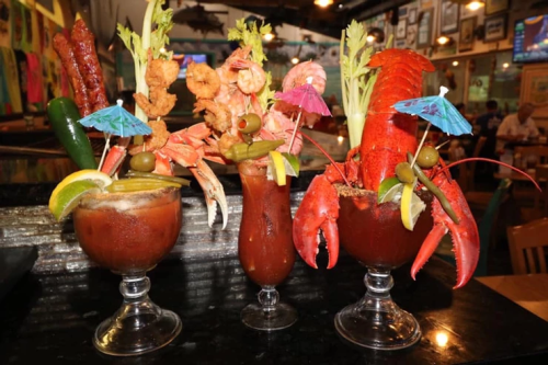 Three colorful seafood cocktails with shrimp, lobster, and garnishes like umbrellas, olives, and lime slices.