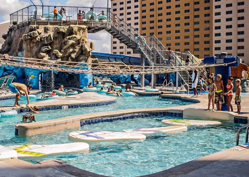 A vibrant water park scene with kids playing, a rock climbing wall, and a water slide in a sunny outdoor setting.