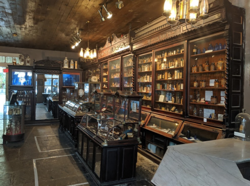A vintage apothecary display with wooden shelves, glass cases, and various jars and bottles in a dimly lit room.