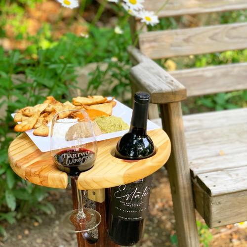 A wooden table holds a glass of wine, a bottle, and a plate of snacks, beside a rustic wooden bench in a garden.