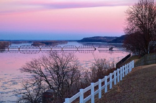 A tranquil river scene at sunset, featuring a bridge, trees, and a white fence against a colorful sky.