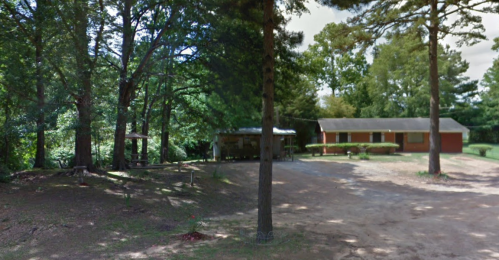 A rural scene featuring a house surrounded by trees and a gravel driveway, with a picnic area nearby.