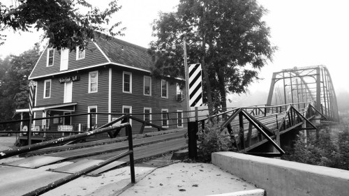 A black and white image of a historic building next to a bridge, surrounded by trees in a foggy setting.