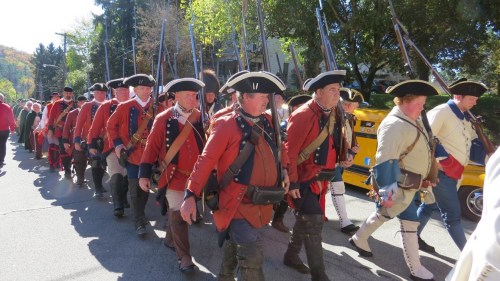 A group of men in colonial military uniforms march in a parade, carrying muskets and wearing tricorn hats.