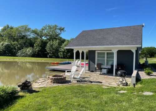 A cozy lakeside cabin with a stone patio, fire pit, and grill, surrounded by greenery and a calm water view.