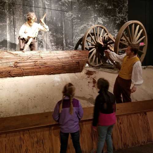 A historical exhibit featuring a figure in a boat, with two children observing and a large wheel in the background.