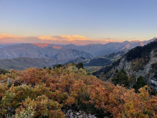 A scenic mountain landscape at sunset, featuring colorful autumn foliage and distant peaks under a pastel sky.