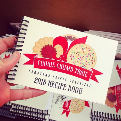 A hand holding a spiral-bound recipe book titled "Cookie Crumb Trail 2018 Recipe Book" with colorful cookie graphics.