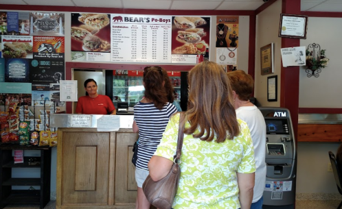 A woman at the counter of Bear's Po-Boys serves customers waiting in line, with a menu displayed above.