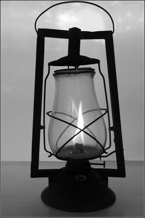 A vintage oil lantern with a visible flame, set against a light background, in black and white.