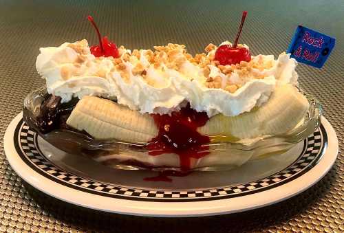 A banana split topped with whipped cream, cherries, nuts, and a "Rock & Roll" flag on a checkered plate.