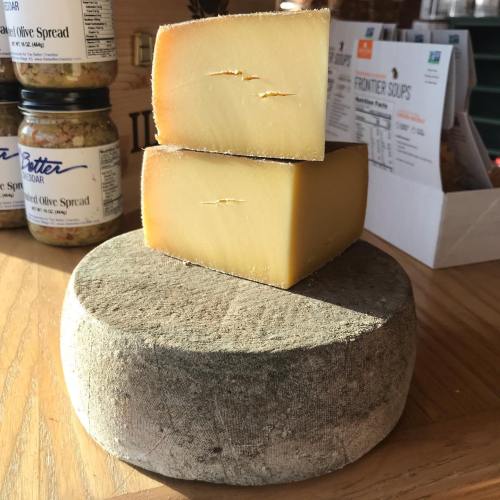 A round wheel of cheese with two wedges cut out, displayed on a wooden surface with jars in the background.