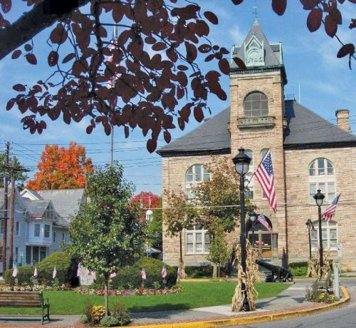 Historic stone building with flags, surrounded by trees and colorful autumn foliage, in a quaint town setting.