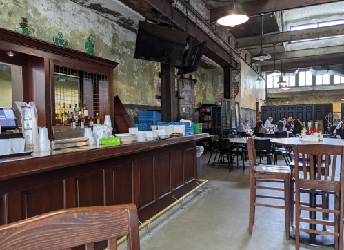 A rustic bar area with wooden furniture, bottles on the counter, and patrons dining in a spacious, industrial-style setting.