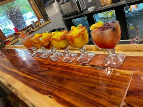 A row of colorful cocktails with fruit garnishes on a wooden bar, with a cooler in the background.