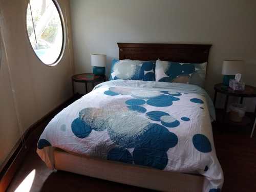 A cozy bedroom featuring a bed with a blue and white patterned quilt, round window, and bedside tables with lamps.