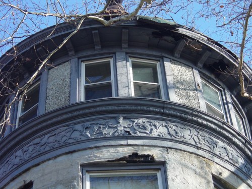 Close-up of a weathered, round turret with ornate detailing and peeling paint, surrounded by bare tree branches.