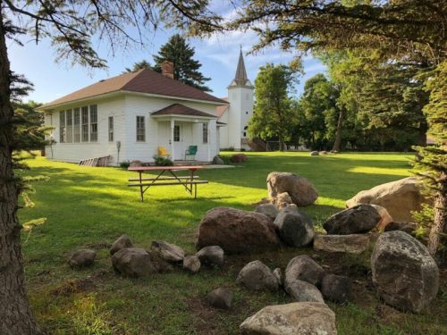 A white house with a red roof and a church steeple in the background, surrounded by green grass and large rocks.