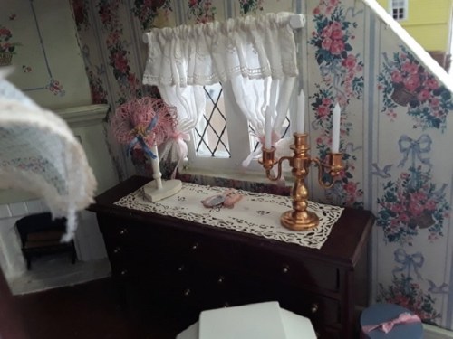 A cozy dollhouse interior featuring a floral wallpaper, a lace curtain, a dresser, and a decorative candle holder.