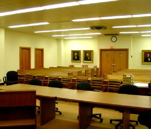 A courtroom interior with wooden tables, chairs, and portraits on the walls, featuring a clock and doors in the background.