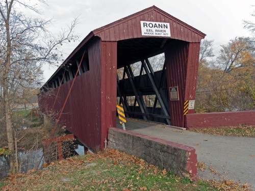 A red covered bridge spans a river, surrounded by trees and autumn foliage, with a sign reading "Roann Eel River Bridge."