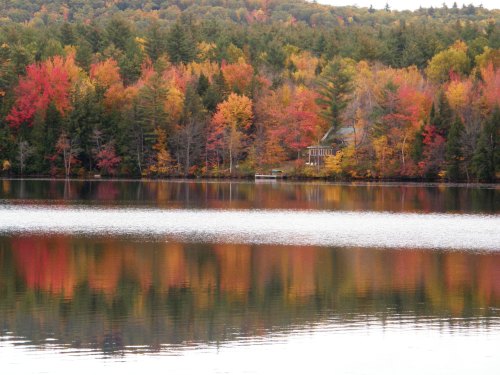 A serene lake reflects vibrant autumn foliage and a cozy cabin surrounded by colorful trees.
