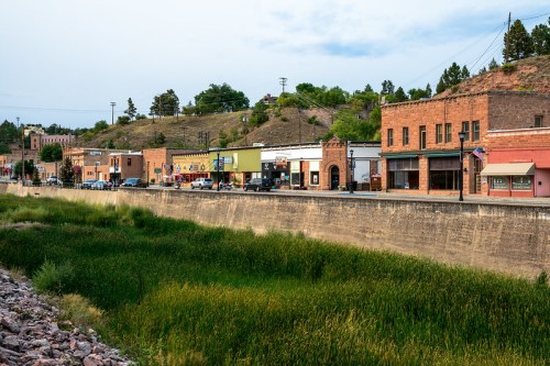 A small town along a river, featuring historic buildings and greenery, with hills in the background under a cloudy sky.