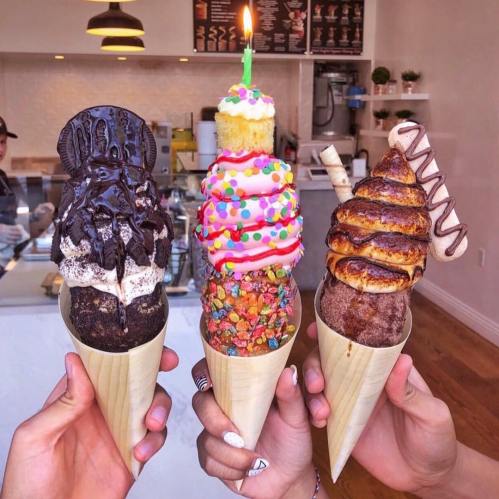 Three colorful ice cream cones with various toppings, including sprinkles and a candle, held by hands in a shop.