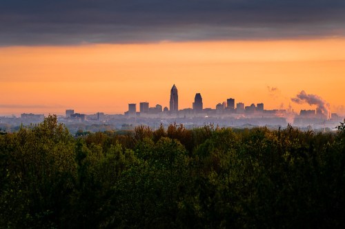 City skyline at sunset, with silhouettes of buildings against a colorful sky, framed by lush green trees in the foreground.