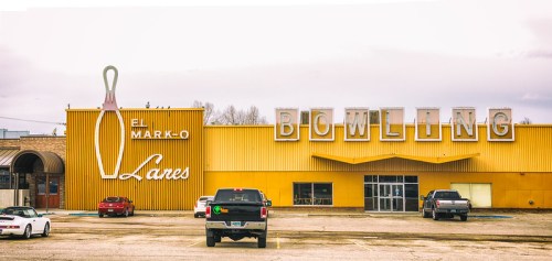 A retro bowling alley with a large sign featuring a bowling pin, surrounded by parked cars.