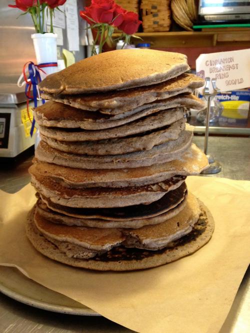 A tall stack of thick pancakes on a plate, with a rustic kitchen background and a small bouquet of roses.