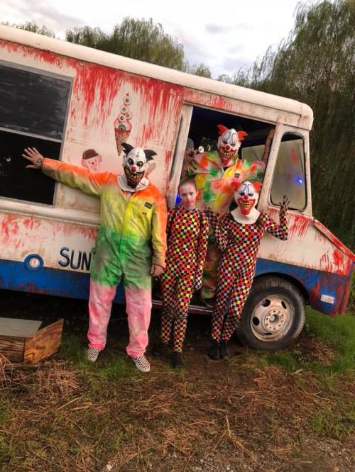 Four clowns in colorful outfits stand by a graffiti-covered ice cream truck, surrounded by trees.