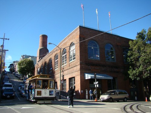 A vintage cable car stops in front of a brick building on a steep street, with clear blue skies above.
