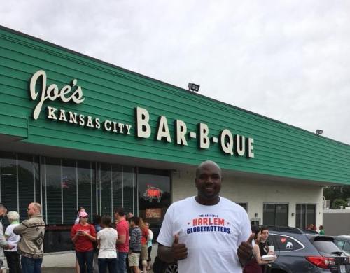 A man in a Harlem Globetrotters shirt stands in front of Joe's Kansas City Bar-B-Que, with a crowd in the background.