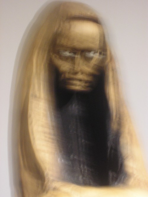 A blurred sculpture of a figure with a solemn expression, carved from wood, set against a plain background.