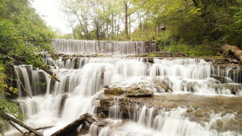 A serene waterfall cascades over rocky ledges, surrounded by lush green trees and a tranquil natural setting.