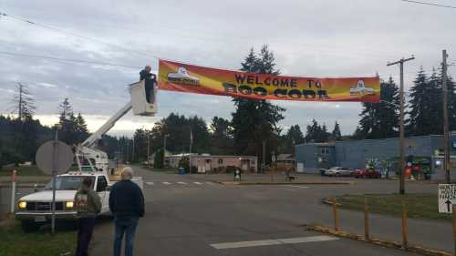 A worker in a lift hangs a "Welcome to Bog Cove" banner over a street, with onlookers nearby. Trees and buildings in the background.