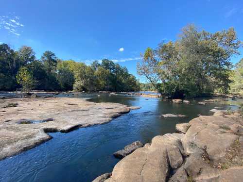 A serene river scene with smooth rocks, trees, and a clear blue sky reflecting in the water.