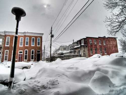 Snow-covered street with buried cars and brick buildings under a gray sky, power lines visible in the background.