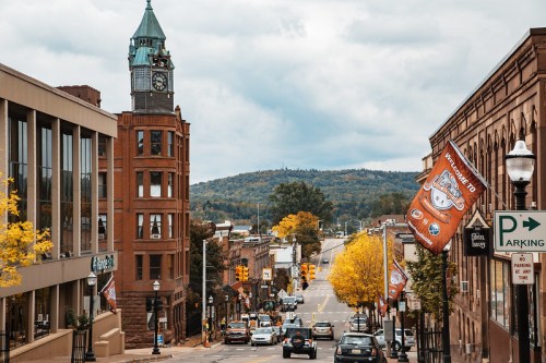 A scenic view of a downtown street with historic buildings, autumn trees, and a clock tower against a cloudy sky.