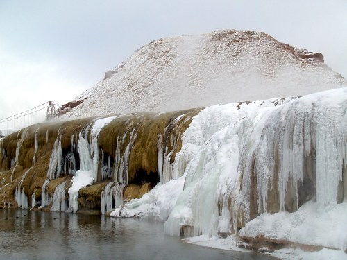 A frozen waterfall cascades over rocky terrain, with steam rising from the water and a snowy hill in the background.