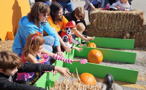 Children and adults push pumpkins down green ramps at a fall festival, surrounded by hay bales and festive decorations.