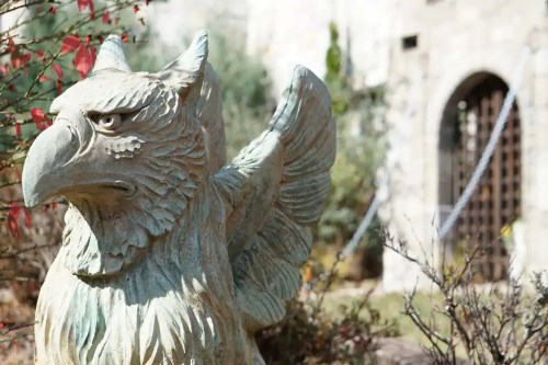 A detailed stone statue of a griffin with wings, surrounded by greenery and a blurred background of a building.