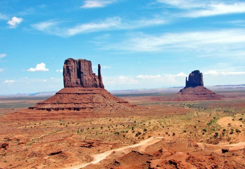 Two iconic rock formations rise from a vast desert landscape under a bright blue sky with scattered clouds.