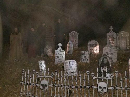 A spooky graveyard scene at night with tombstones, a fence with skulls, and shadowy figures in the background.