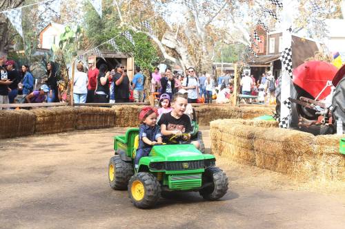 Two children ride a toy tractor through a festive outdoor area filled with people and hay bales.