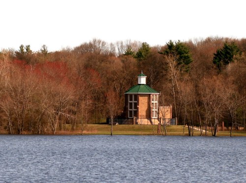 A brick building with a green roof stands by a calm lake, surrounded by trees and a clear sky.