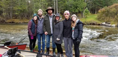A group of six people stands by a river, surrounded by trees, smiling and dressed for outdoor activities.