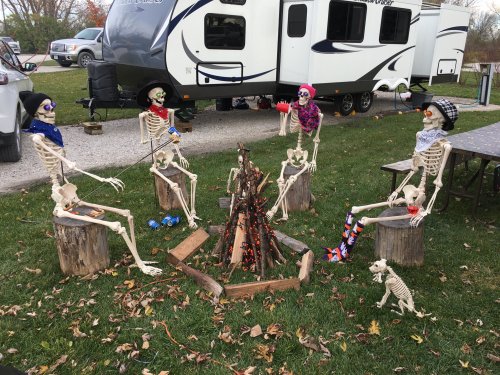 Five skeletons sitting around a campfire, wearing hats and scarves, with a camper in the background.