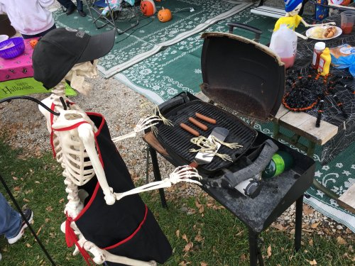 A skeleton in an apron and cap grills hot dogs at a festive outdoor gathering, surrounded by Halloween decorations.